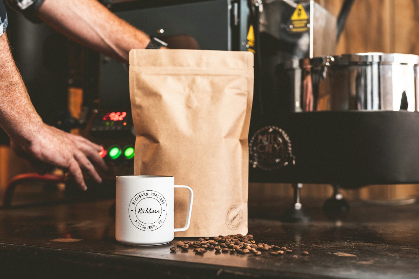 Hender som betjener en kaffebrenner nær bønner og en kopp.